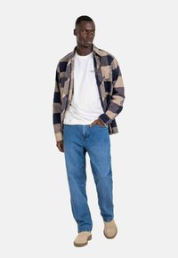 Camisa de franela a cuadros en beige y azul marino, camiseta blanca debajo, jeans azules y calzado de color marrón claro, de pie contra un fondo neutro.