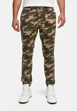 Homme portant un pantalon de jogging camouflage vert et marron avec des poignets élastiques et des baskets blanches montantes.