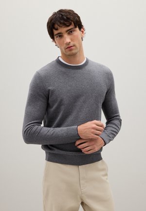 Pull en maille gris avec col rond et poignets côtelés, porté sur une chemise blanche. Le tissu semble doux et texturé, style ajusté.