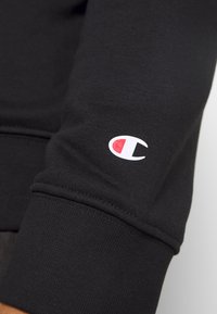 Sort sweatshirt stof med ribbede manchetter, prydet med et hvidt logo med et rødt accent på venstre ærme. Nærbillede af teksturen.