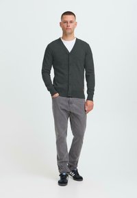 Bărbatul stă cu o mână în buzunar, purtând un cardigan cu nasturi de culoare gri închis, o cămașă albă, pantaloni gri și adidași negri pe un fundal alb.