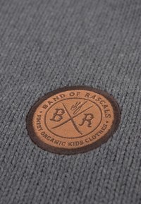 Grå strikket stof med en brun cirkulær lap med præget logo og tekst, der angiver mærke og økologisk materiale, nederst til højre.