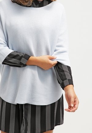 Persona ajustando el puño de un suéter azul claro acanalado, llevado sobre un vestido negro con rayas verticales blancas.