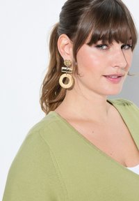 Guldtonade örhängen med en texturerad rund topp och en stor vävd rund ring. Toppdelen har en glänsande yta, och ringen är ljusbeige.