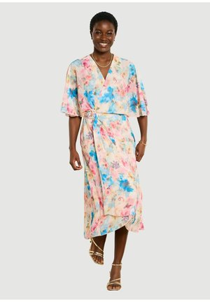 Usmievavá žena v pastelovom kvetovanom zavinovacom šate s širokými rukávmi a zlatých sandáloch na remienky, kráčajúca na svetlom jednoduchom pozadí.