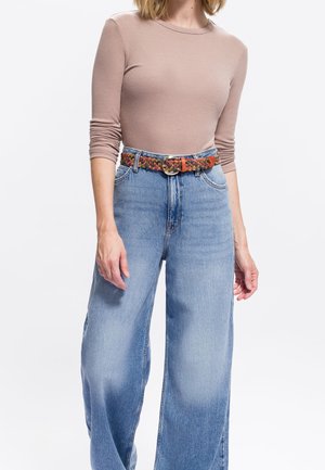 Woman wearing a fitted long-sleeve beige top tucked into high-waisted wide-leg blue jeans with a braided brown belt.