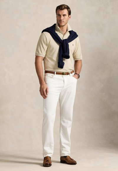 Mies seisoo beige polo päällään, valkoiset housut, ruskea vyö ja kengät, tummansininen neule olkapäillä yksiväristä taustaa vasten.