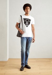 Camiseta blanca con un gráfico de pantera negra, combinada con jeans de mezclilla azul claro y mocasines negros. El modelo sostiene un bolso mensajero oscuro.