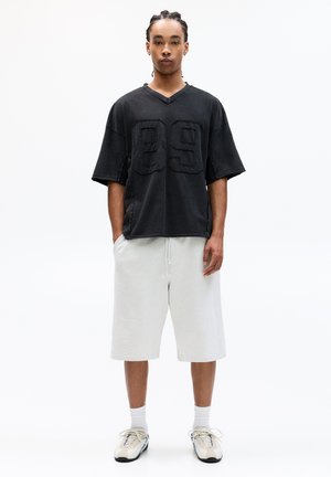 Černé oversized tričko s designem "89", spárované se světle šedými šortkami. Tkanina vypadá měkce a oblečení doplňují sportovní boty.