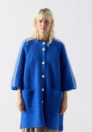 Femme aux cheveux blonds portant un cardigan bleu ample boutonné avec des rayures blanches sur les manches et de grandes poches avant, debout devant un fond uni.