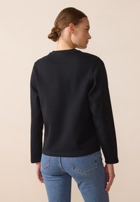 Svart hettegenser med lange ermer, rund halslinning og minimal søm. Kombinert med lyseblå tettsittende jeans. Glatt stoffstruktur.