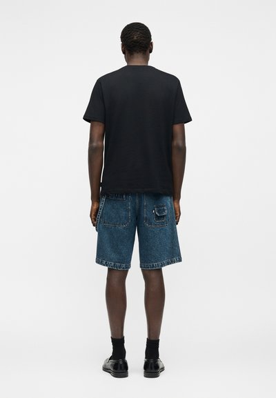 Άνδρας που στέκεται με την πλάτη, φορώντας μαύρο μπλουζάκι, μπλε τζιν σορτς με πίσω τσέπες, μαύρες κάλτσες και μαύρα παπούτσια μπροστά σε απλό φόντο.