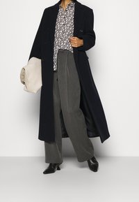 Cappotto di lana blu navy, camicia a righe con bottoni, pantaloni grigi e stivaletti neri. Borsa intrecciata beige tenuta sotto il braccio.