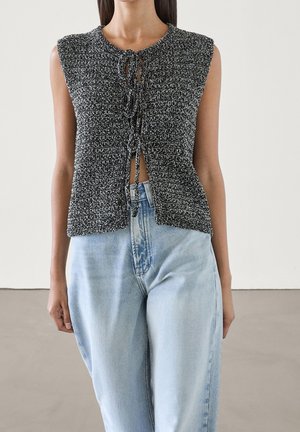 Woman wearing sleeveless black and white knit top with front ties paired with light blue high-waisted jeans against plain background.