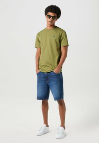 Zielona bawełniana koszulka zestawiona z niebieskimi dżinsowymi szortami, białymi tenisówkami i ciemnymi okularami przeciwsłonecznymi. Prosty design z logo na koszulce.