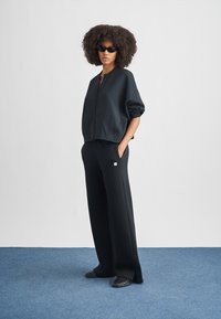 Sweatshirt preta com fecho de correr e mangas curtas, combinada com calças pretas largas. Modelo usa óculos de sol pretos e sapatos rasos.