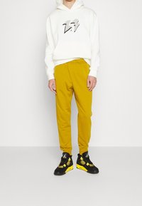 Felpa bianca con grafica a saetta nera, abbinata a pantaloni da tuta giallo senape e sneakers nere con accenti gialli.