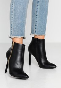 Stivaletti neri in pelle con punta affusolata e tacco a stiletto. Caratteristiche includono zip laterali e una texture liscia. Indossati con jeans a fondo sfrangiato.