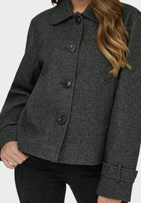 Šedý kabát vzoru houndstooth vyrobený z texturované tkaniny, s límcem a čtyřmi černými knoflíky vpředu.