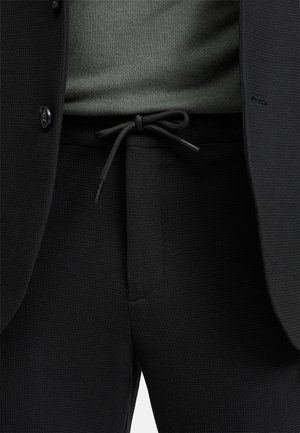 Svarte teksturerte bukser med snor i livet, kombinert med en mørkegrønn topp og en svart blazer. Har knapper og en glatt stoff.