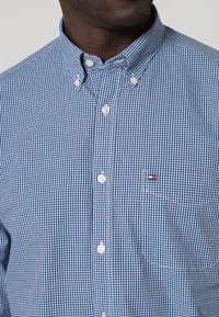 Camisa abotonada en patrón de cuadros azul marino y blanco, con cuello estándar, botones blancos y un pequeño logo en el bolsillo del pecho.