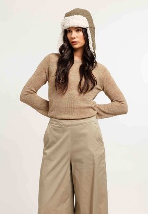 Femme aux longs cheveux noirs portant un pull beige, un pantalon large en kaki et un bonnet d'hiver brun avec une doublure en polaire blanche.