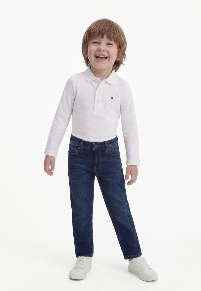 Osmijeh mladi dječak svijetlosmeđe kose, odjeven u bijeli polo s dugim rukavima, tamnoplave traperice i bijele tenisice, stoji na bijeloj pozadini.