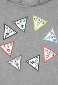 Sudadera gris con ocho parches triangulares de varios colores, cada uno de ellos con el texto "GUESS U.S.A." y un signo de pregunta.