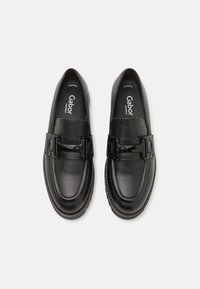Mocassins en cuir noir avec une boucle noire brillante, texture lisse, bout arrondi et accent cousu autour de la semelle. Logo de la marque visible à l'intérieur.