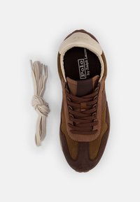 Brun sneaker med mocka och meshmaterial, rund tå och beige vadderad krage. Inkluderar ljusbruna snören bredvid skon.