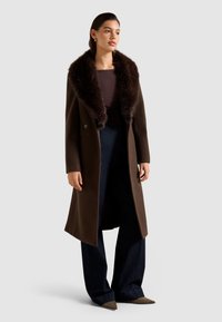 Abrigo de lana marrón con un gran cuello de piel oscura, diseño de doble botonadura, combinado con pantalones de mezclilla oscuros de pierna ancha y zapatos de punta.