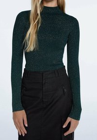 Groene top met lange mouwen en hoge hals, gemaakt van ribstof met metalen draden; gecombineerd met zwarte cargobroek met ritsen.