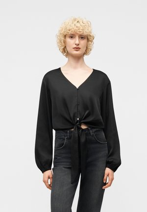 Lyshåret kvinde med krøllet hår iført en sort langærmet knappet bluse bundet i taljen og mørke high-waist jeans.