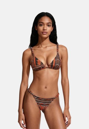 Donna con capelli lunghi e ricci che indossa un bikini due pezzi marrone e arancione con fantasia, su uno sfondo bianco uniforme.