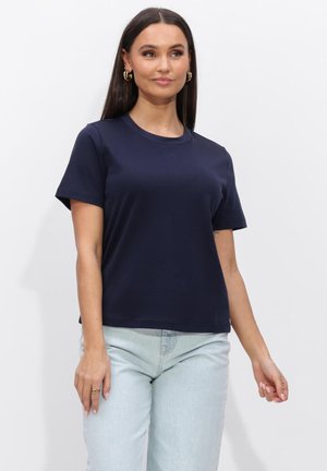 Vrouw met lang donker haar, goudkleurige creolen, een marineblauw t-shirt met korte mouwen en lichtblauwe spijkerbroek, staand tegen een witte achtergrond.