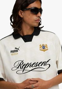 Maglia bianca a motivo con collo nero e dettagli logo. Presenta la scritta "Represent" in corsivo nero. Modello con occhiali da sole, braccia incrociate.