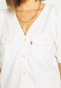 Chemise en coton blanc avec col en V, manches courtes bouffantes, deux poches poitrine et boutons en perle. Comprend une petite étiquette logo Levi's rouge.