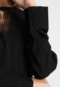 Blusa nera con scollatura in pizzo, maniche voluminose con bottoni alle estremità e una texture liscia e opaca.