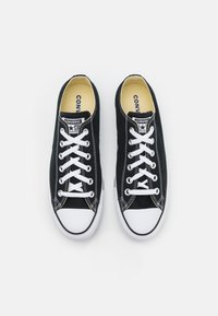 Schwarze Leinenschuhe mit weißen Schnürsenkeln, Gummikappe und kontrastierenden weißen Nähten. Das Innenfutter verfügt über eine gelbe Einlegesohle mit Branding.