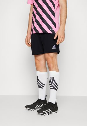 Mężczyzna w czarnych szortach Adidas, różowo-czarnej koszulce w paski, białych wysokich skarpetach z czarnymi wzorami oraz czarnych korkach piłkarskich Adidas.