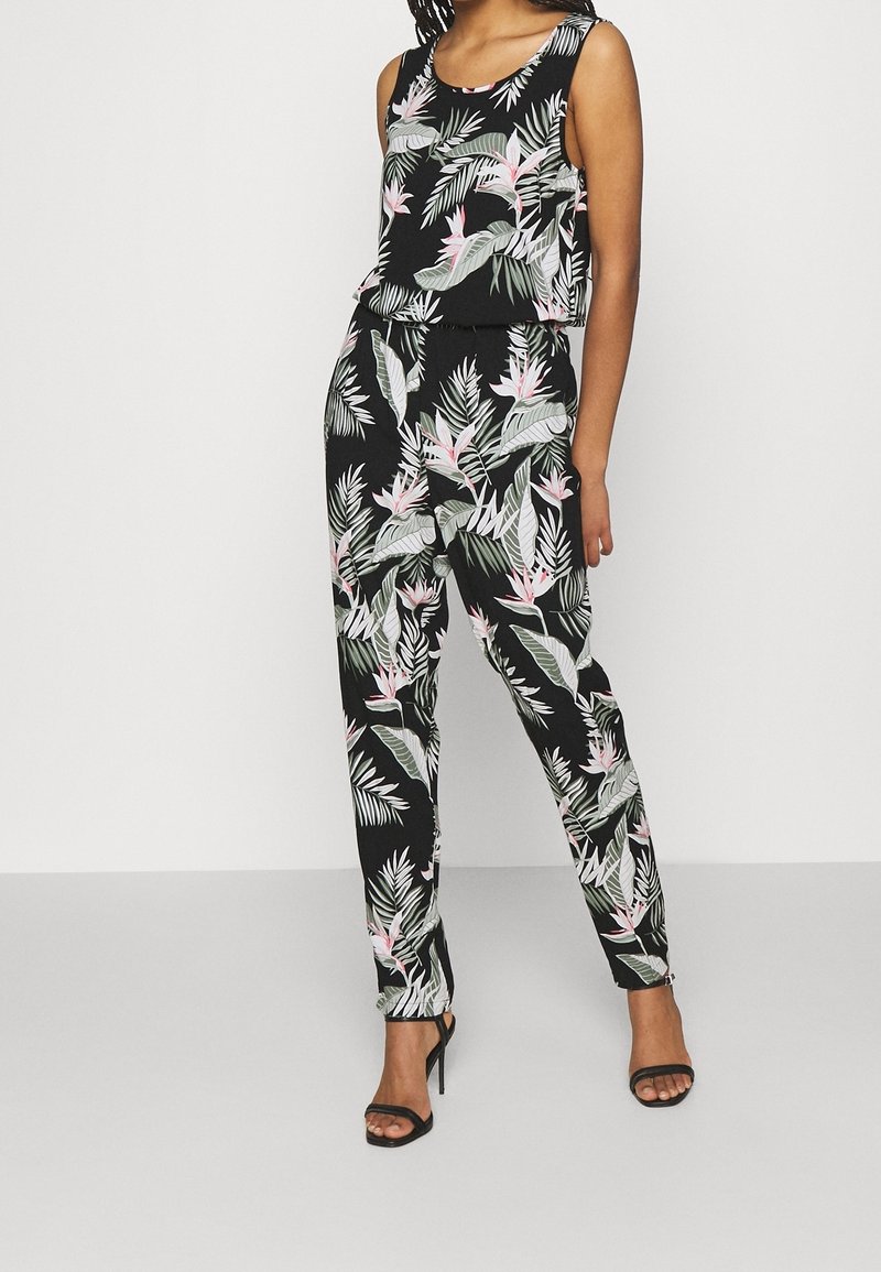 Femme portant une combinaison sans manches noire à motifs floraux avec des feuilles roses et vertes, associée à des sandales noires à talons hauts, posant devant un fond blanc.