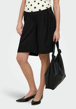 Nő fekete, térdig érő rövidnadrágot és fekete hegyes lapos cipőt visel, kezében fekete bőrtáskával, fehér pöttyös felsővel.