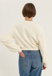Felpa corta di colore crema chiaro con maniche lunghe e leggermente a sbuffo, orlo elastico e cuciture in vista. Indossata con jeans blu a vita alta.