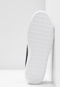 Semelle d'une basket blanche Puma avec un motif étoile texturé et le logo Puma au talon, placée verticalement contre des murs d'angle blancs.
