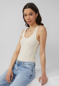 Crème tanktop van geribbeld materiaal met een ronde halslijn. Gecombineerd met lichtblauwe, hoge taille jeans, zittend op een witte ondergrond.