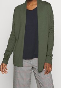 Öppen cardigan i olivgrön tyg med långa ärmar, lager på lager över en marinblå V-ringad topp och i kombination med rutiga byxor med röda accenter.