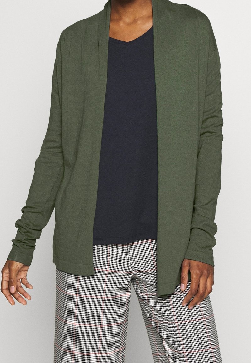 Öppen cardigan i olivgrön tyg med långa ärmar, lager på lager över en marinblå V-ringad topp och i kombination med rutiga byxor med röda accenter.