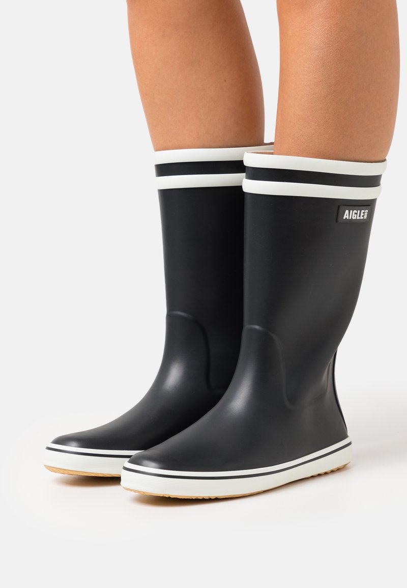 Bottes de pluie en caoutchouc noires avec une finition texturée, rayures blanches en haut et une semelle en caoutchouc beige contrastante. Logo visible sur le côté.