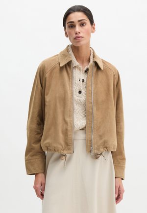 Frau trägt eine beige Wildlederjacke mit Reißverschluss über einem cremefarbenen Strickpullover und einem hellbeigen Rock, steht vor einem schlichten weißen Hintergrund.