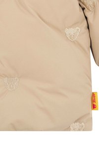 Chaqueta acolchada beige con motivos de osos bordados, textura suave y una etiqueta de Steiff amarilla y roja en la parte inferior.